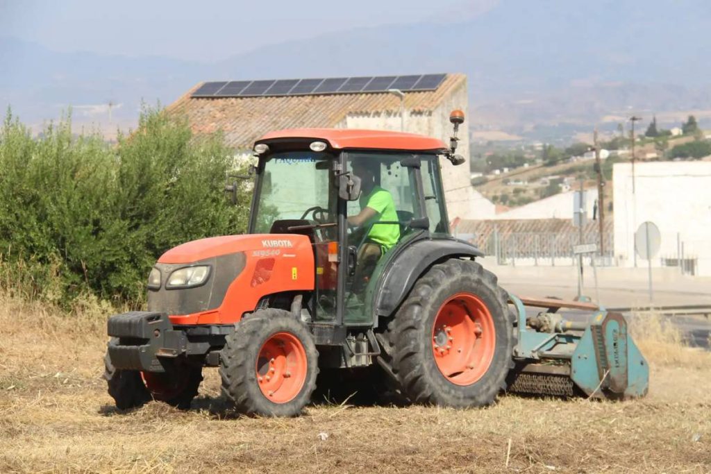 Un tractor con desbrozadora retira la hierba seca de una parcela en Cártama, con el pueblo y las montañas al fondo